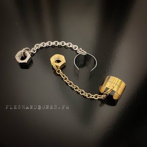 Könnte beinhalten: Eine goldene und silberne Kette mit zwei Muttern daran. Die Kette ist mit einem goldenen Ring verbunden, auf dem der Text "FLESH AND BONES" eingraviert ist. Der Ring ist offen und kann am Finger getragen werden.