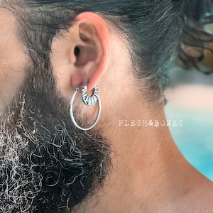 May include: A silver hoop earring with a textured design. The earring is worn in the left ear of a person with a beard.
