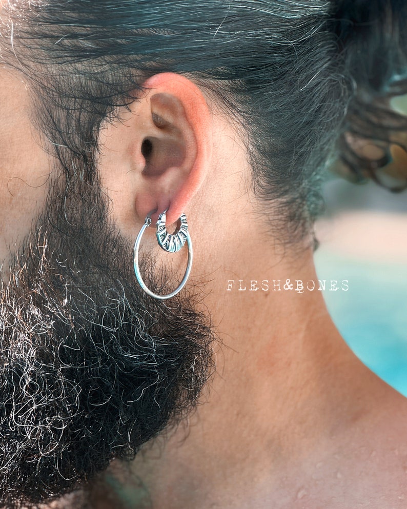 May include: A silver hoop earring with a textured design. The earring is worn in the left ear of a person with a beard.