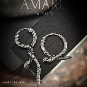 Puede incluir: Dos pendientes de aro de serpiente plateados, cada uno con un diseño de serpiente detallado. Los pendientes son parte de un paquete dúo, como lo indica el texto "DUO PACK" en el fondo negro. El nombre de la marca "AMARU" también es visible en el fondo.