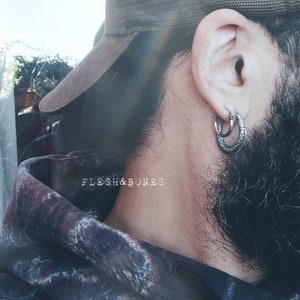 Puede incluir: Una persona con una gorra de b&eacute;isbol de malla marr&oacute;n con un aro de plata en la oreja izquierda. La persona tiene barba y lleva una sudadera con capucha azul oscuro con un estampado floral.