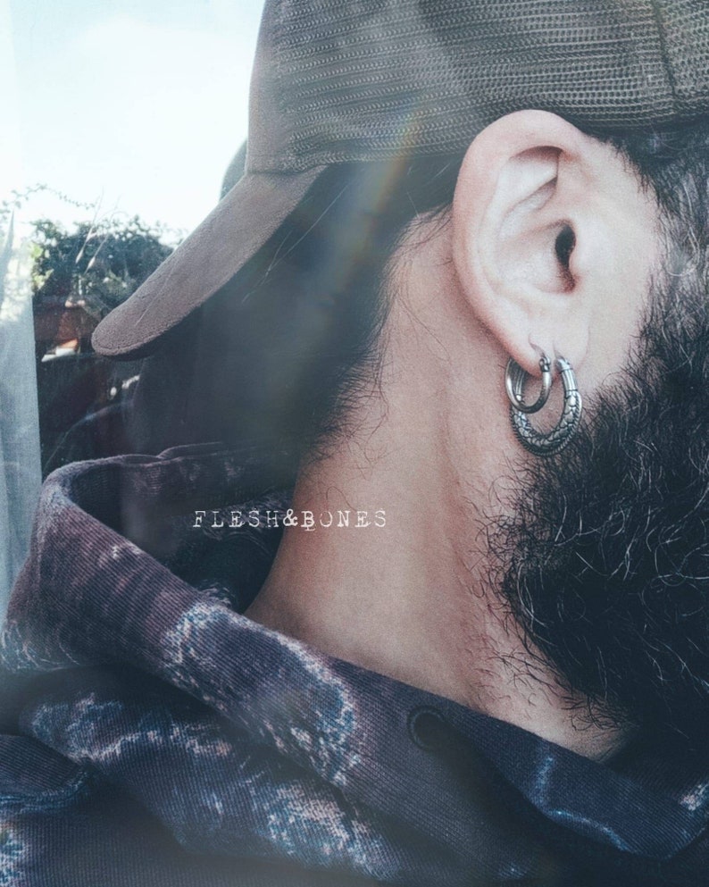 Puede incluir: Una persona con una gorra de b&eacute;isbol de malla marr&oacute;n con un aro de plata en la oreja izquierda. La persona tiene barba y lleva una sudadera con capucha azul oscuro con un estampado floral.