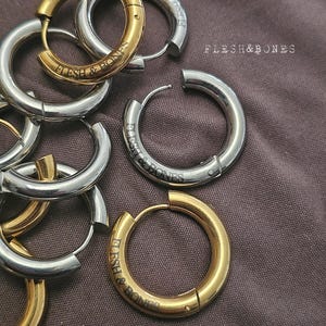 Peut inclure: Plusieurs boucles d'oreilles créoles dorées et argentées. Chaque boucle d'oreille porte l'inscription "FLESH & BONES". Les boucles d'oreilles sont posées sur un tissu marron.