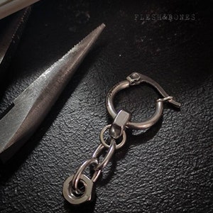 May include: A silver hoop earring with a small chain and a nut charm. The earring is on a black surface with the text "FLESH&BONES" visible in the background.