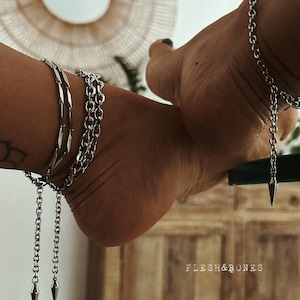 Könnte beinhalten: Silberfarbene Fußkettchen mit hängenden, konisch geformten Anhängern. Die Fußkettchen haben ein Kettenmuster und werden an den Knöcheln getragen. Das Bild zeigt auch den Text "FLESH&BONES".