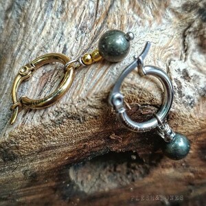 May include: A pair of earrings with gold and silver hoops and green gemstone beads. The earrings are on a wooden surface.