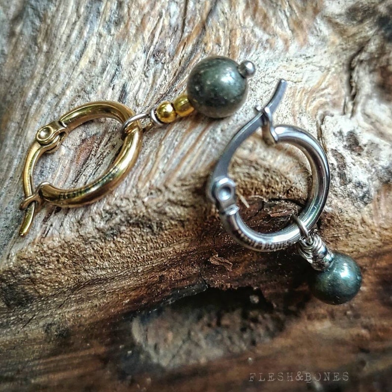 May include: A pair of earrings with gold and silver hoops and green gemstone beads. The earrings are on a wooden surface.