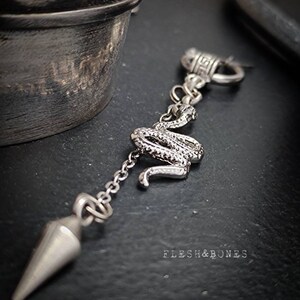 Puede incluir: Un colgante de plata con un dije de serpiente, una cadena y un cono puntiagudo. El colgante cuelga de un anillo de plata con un dise&ntilde;o de llave griega. El texto "FLESH & BONES" es visible en la esquina inferior derecha de la imagen.