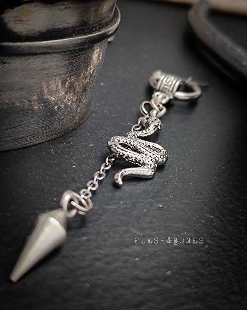 Puede incluir: Un colgante de plata con un dije de serpiente, una cadena y un cono puntiagudo. El colgante cuelga de un anillo de plata con un dise&ntilde;o de llave griega. El texto "FLESH & BONES" es visible en la esquina inferior derecha de la imagen.