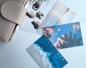 May include: A beige planner with a pen and several decorative inserts. The inserts feature images of seashells, ocean waves, and tropical skies with palm trees. The planner and inserts are on a white surface.