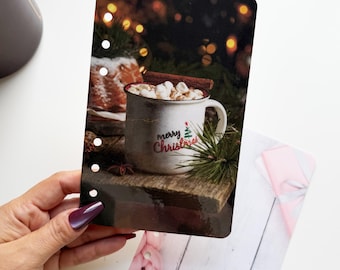 May include: A decorative card featuring a cozy Christmas scene. A mug of hot chocolate with marshmallows and a cinnamon stick sits on a wooden surface. The mug has the words "merry Christmas" and a Christmas tree. The background has blurred lights.