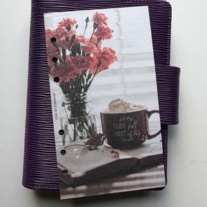 May include: A dark purple planner with a textured cover featuring a vertical insert. The insert displays a glass vase with red carnations, a mug with whipped cream and the text I LOVE fall MOST of ALL, and an open book. AHORROS DE NEDI is printed.