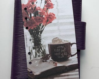 May include: A dark purple planner with a textured cover featuring a vertical insert. The insert displays a glass vase with red carnations, a mug with whipped cream and the text I LOVE fall MOST of ALL, and an open book. AHORROS DE NEDI is printed.