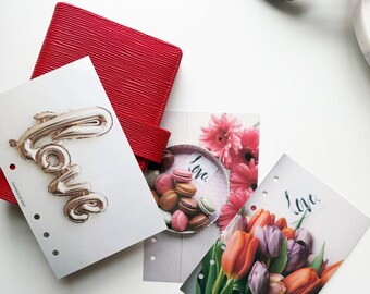 May include: A red textured planner with several decorative inserts. One insert features a gold balloon spelling "Love", another shows macarons, and a third displays a bouquet of tulips. The inserts are on a white surface.