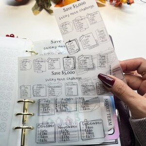 以下が含まれることがあります： 手書きのテキストが入った透明な付箋チャレンジシート。貯蓄を追跡するように設計されています。シートはバインダーに保持され、「Save 5.000 $ Sticky note challenge」というテキストが表示されます。各月には貯蓄目標があります。