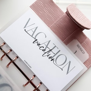 May include: A light pink planner with a textured cover and rose gold rings. A white insert displays the word "VACATION" in gray and black script. A small tab reads "MEXICO". The planner is open, revealing the insert.