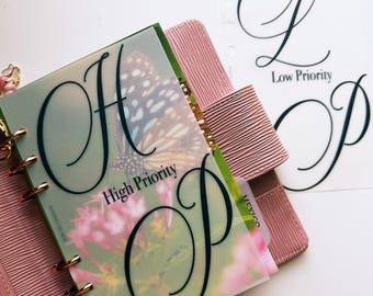 May include: A pink textured ring binder planner containing a translucent divider with a butterfly and flower print, featuring the black cursive letters H and P and the text High Priority, alongside a separate white sheet with the letters L and P.