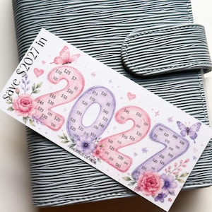 May include: A white rectangular savings challenge card with the text Save $2027 in 2027 and AHORROS DE NEDI. The card features large pink and purple numbers filled with dollar amounts, decorated with watercolor butterflies, hearts, and floral accents.