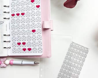 May include: A pink planner with a money-saving tracker titled "Money Goals: One Envelope at a Time." The tracker features numbered envelope illustrations, some with pink hearts. A pen and sticker sheet with the same design are also visible.