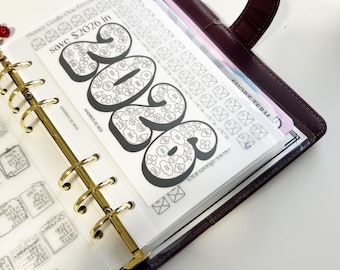 May include: A burgundy leather planner with gold-colored rings, open to a page with a savings tracker. The tracker is a black and white design with the year "2026" in large numbers, and the text "Money Goals One envelope at a time save $2026 in".