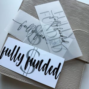 以下が含まれることがあります： 黒いスクリプト体と太字でfully fundedと書かれた3枚の長方形ステッカー。2枚は半透明で黒い文字、1枚は不透明な白地に黒い文字が印刷されています。各ステッカーには大きな灰色のドル記号が表示されています。