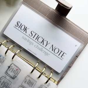 May include: A beige planner with gold rings and a clear pocket holding a white card. The card reads "$10K STICKY NOTE savings challenge" in black text. The planner is open, revealing pages with handwritten notes.