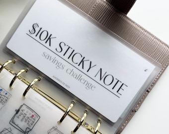 May include: A beige planner with gold rings and a clear pocket holding a white card. The card reads "$10K STICKY NOTE savings challenge" in black text. The planner is open, revealing pages with handwritten notes.