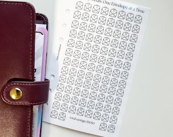 May include: A burgundy leather planner with a gold button clasp next to a white paper with a savings tracker. The paper has the text "Money Goals: One Envelope at a Time" and a grid of numbered envelopes.