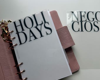 May include: A pink textured planner with gold rings and a translucent divider featuring the black text HOLI DAYS. A separate translucent sheet with the black text NEGOCIOS is positioned to the right. The planner includes a gold charm attachment.