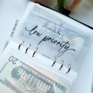 Puede incluir: Una página de plástico transparente con el texto "low priority" escrito en cursiva negra. La página está diseñada para ser utilizada en una carpeta o planificador. Se muestra con billetes de banco en su interior.