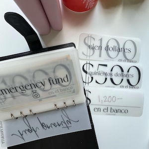 以下が含まれることがあります： 「emergency fund」と書かれた透明ポケットが付いた黒いバインダー。その横には、スペイン語でドル金額が書かれた白いラベルが並んでいます。100ドル、500ドル、1,200ドルで、すべて「en el banco」と表示されています。