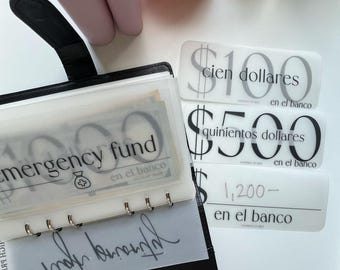 May include: A black binder with clear pockets, one labeled "emergency fund." Beside it are white labels with dollar amounts in Spanish: $100, $500, and $1,200, all marked "en el banco."