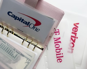 May include: A light pink textured binder with gold rings containing a Capital One card and a five-dollar bill in a clear sleeve. Beside the binder are two translucent dividers with the printed text T-Mobile and Verizon in pink and red.