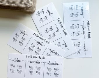 May include: White cards with black text, designed for tracking savings. Each card is labeled with a month and has spaces to record amounts. The text "I will save $20k" is written on each card. A beige wallet is in the background.
