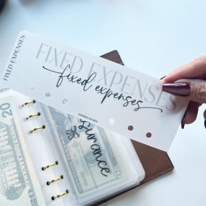 以下が含まれることがあります： 「FIXED EXPENSES」の文字がグレーと黒の筆記体で印刷された白いプランナーディバイダー。ディバイダーは、現金が入った透明なプランナーポケットの上に保持されています。プランナーは金色のリングと茶色のカバーが付いています。