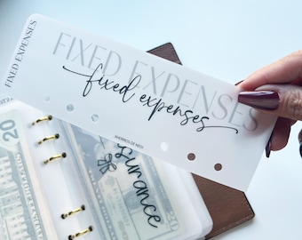 May include: A white planner divider with the words "FIXED EXPENSES" printed in gray and black script. The divider is held over a clear planner pocket containing cash. The planner has gold rings and a brown cover.