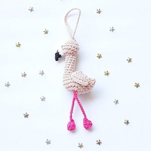 May include: A pink crocheted flamingo ornament with black eyes and pink legs. The flamingo is hanging from a white string.