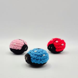 May include: Three crocheted ladybugs in pink, blue, and red with white spots and black accents. The ladybugs are small and round, with a detailed crochet pattern. The background is plain white.