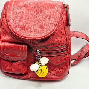 May include: A bright red leather backpack with a textured surface, featuring multiple zippered compartments and a silver-toned metal zipper pull. A small, handmade yellow, black, and white crocheted bee charm is attached to the front zipper.
