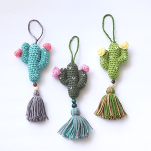 May include: Three crocheted cactus ornaments with tassels. The cacti are green with pink and yellow flowers. The tassels are gray, green, and blue.