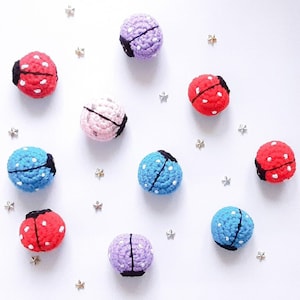May include: A collection of ten crocheted ladybug ornaments in various colors, including red, blue, pink, and purple. The ornaments are arranged on a white background with silver glitter stars.