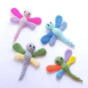 May include: Four colorful, crocheted dragonfly toys. Each dragonfly has a round head, a slender body, and four wings. The wings are various colors, including pink, blue, green, yellow, and orange. The dragonflies are arranged on a white surface.