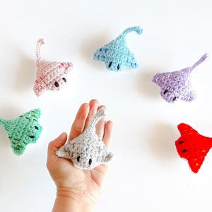 May include: Six crocheted stingray plushies in various colors, including pink, blue, purple, green, gray, and red. The plushies have black eyes and are arranged on a white surface.