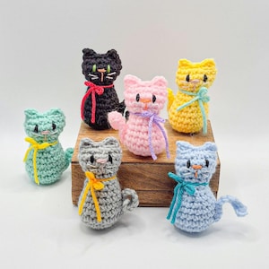 May include: A collection of six crocheted cat figurines in various colors, including black, pink, yellow, blue, gray, and teal. Each cat has a contrasting ribbon bow and embroidered facial features. The cats are displayed on a wooden block.