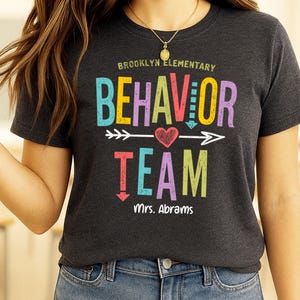 May include: Dark gray t-shirt with the words "Brooklyn Elementary BEHAVIOR TEAM" in colorful letters. A heart and arrow design is between "BEHAVIOR" and "TEAM". The name "Mrs. Abrams" is printed below.