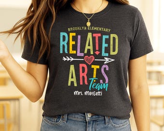May include: Dark gray t-shirt with the text "BROOKLYN ELEMENTARY RELATED ARTS TEAM" in colorful block letters. A white arrow with a heart points to the word "ARTS". The name "Mrs. Massetti" is below the text.