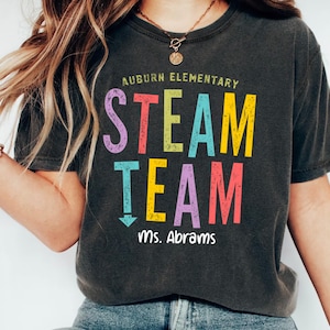 May include: Dark gray t-shirt with the words "Auburn Elementary STEAM TEAM Ms. Abrams" in colorful block letters. The shirt is a casual style, suitable for teachers or students. The text is in various colors, including pink, yellow, blue, and purple.