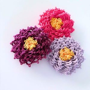 May include: Three crocheted flowers in shades of pink, purple, and yellow. The flowers are layered and have a delicate, intricate design.