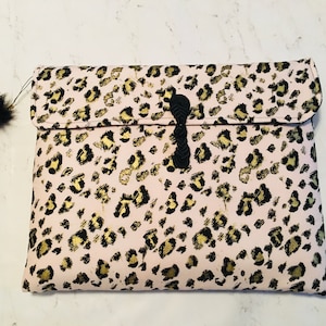 Puede incluir: Un clutch rosa con un estampado de leopardo en negro y dorado. El clutch tiene un cierre de cuerda negra y un pompón negro y esponjoso adjunto. La solapa está doblada y asegurada con una cuerda negra.