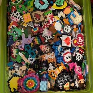 Op de afbeelding: Een groene plastic container gevuld met kleurrijke pixelkunst gemaakt van kleine plastic kralen. De pixelkunst omvat verschillende ontwerpen, zoals harten, sterren en cartoonfiguren.